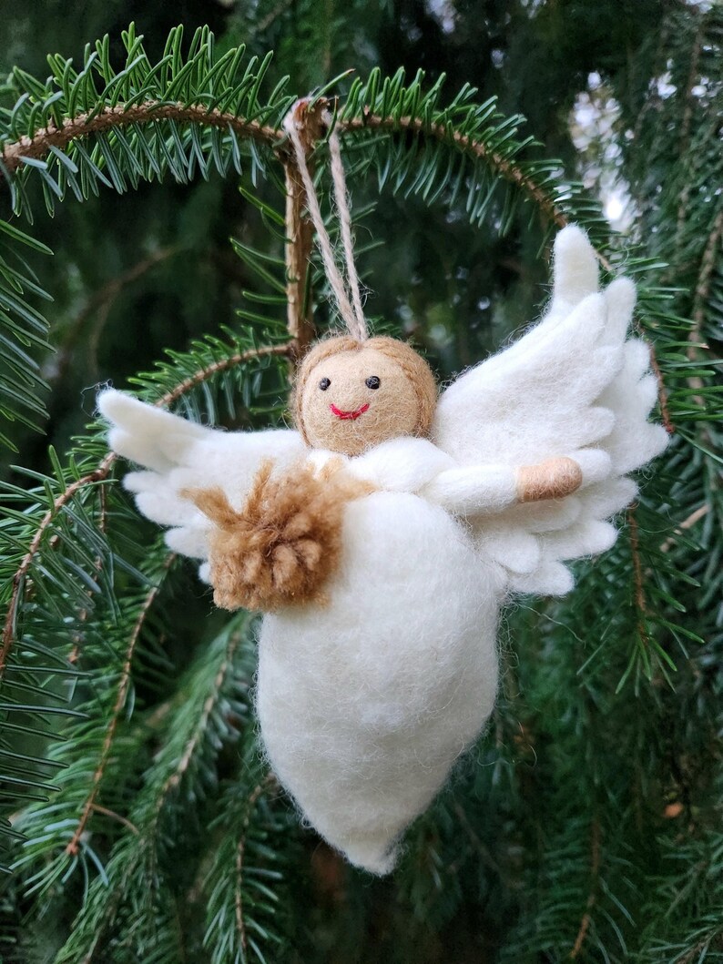 Puede incluir: Un adorno de &aacute;ngel de fieltro blanco con alas de fieltro marr&oacute;n y una cara sonriente. El &aacute;ngel sostiene una bola de fieltro marr&oacute;n en sus manos.