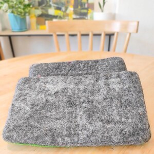 May include: Two rectangular felted wool pads in a mottled charcoal gray color. The pads have a thick, dense, and textured surface with visible fiber strands. They are stacked slightly offset, revealing a thin, vibrant green layer at the base.