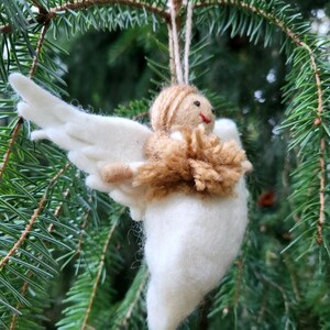 Puede incluir: Un adorno de &aacute;ngel de fieltro blanco con pelo de lana marr&oacute;n y un vestido de lana marr&oacute;n. El &aacute;ngel tiene una cara sonriente y alas.