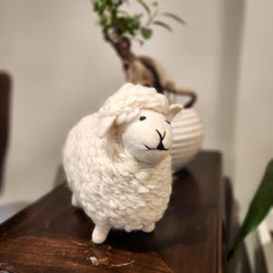 May include: A handcrafted, off-white sheep figurine with a fluffy wool texture. The sheep has black eyes and a black 'Y' shaped mouth. The figurine is a decorative item, possibly for a nursery or farmhouse decor.