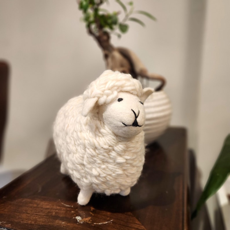 May include: A handcrafted, off-white sheep figurine with a fluffy wool texture. The sheep has black eyes and a black 'Y' shaped mouth. The figurine is a decorative item, possibly for a nursery or farmhouse decor.