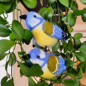 May include: Two bluebirds with yellow bellies and green wings are perched on a green leafy plant. The birds are made of felt and have a hand-crafted appearance.