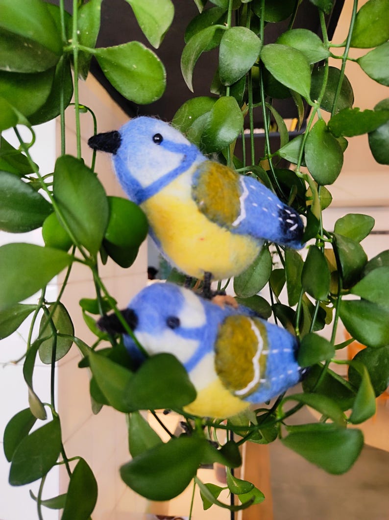 May include: Two bluebirds with yellow bellies and green wings are perched on a green leafy plant. The birds are made of felt and have a hand-crafted appearance.