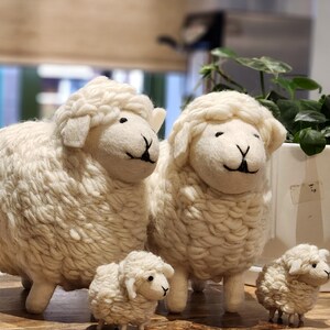 May include: Four white wool sheep figurines of varying sizes. The sheep have black eyes and a stitched black mouth. The wool is textured and fluffy. The sheep are standing on a wooden surface.