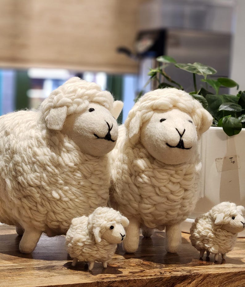 May include: Four white wool sheep figurines of varying sizes. The sheep have black eyes and a stitched black mouth. The wool is textured and fluffy. The sheep are standing on a wooden surface.