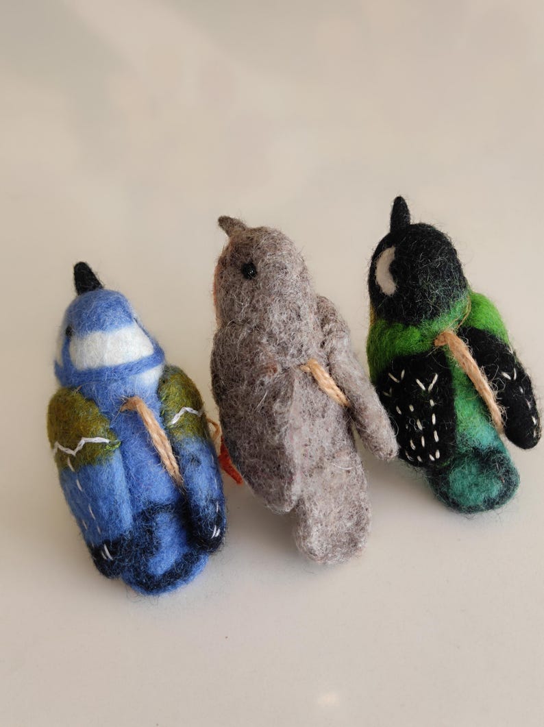 May include: Three felt bird ornaments, each with a different colour and pattern. The birds are blue, grey, and green with black accents. They are all tied together with twine.