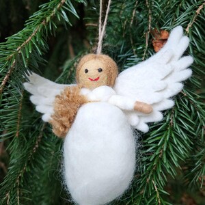 Puede incluir: Un adorno de &aacute;ngel de fieltro blanco con pelo y alas de fieltro marr&oacute;n. El &aacute;ngel tiene una cara sonriente y est&aacute; colgado de una cuerda.