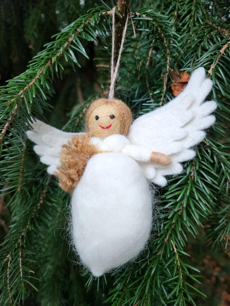 Puede incluir: Un adorno de &aacute;ngel de fieltro blanco con pelo y alas de fieltro marr&oacute;n. El &aacute;ngel tiene una cara sonriente y est&aacute; colgado de una cuerda.