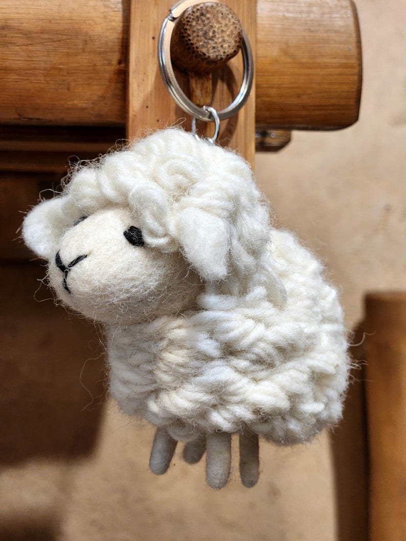 May include: A white sheep keychain made of textured yarn. The sheep has a fluffy body, small black eyes, and a black stitched mouth. The keychain is attached to a silver metal ring.