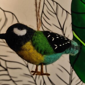 May include: A felt bird ornament in black, white, green, yellow and blue colours. The bird has a small black beak and white markings on its head.