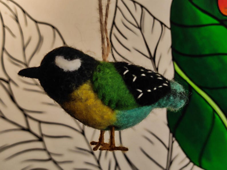 May include: A felt bird ornament in black, white, green, yellow and blue colours. The bird has a small black beak and white markings on its head.