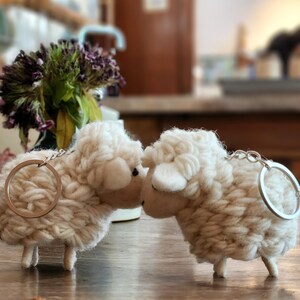 May include: Two off-white sheep keychains made of a textured, wool-like material. Each sheep has a silver-colored keyring attached. The sheep are positioned close together, suggesting a pair. The background is blurred, with hints of a floral arrangement.