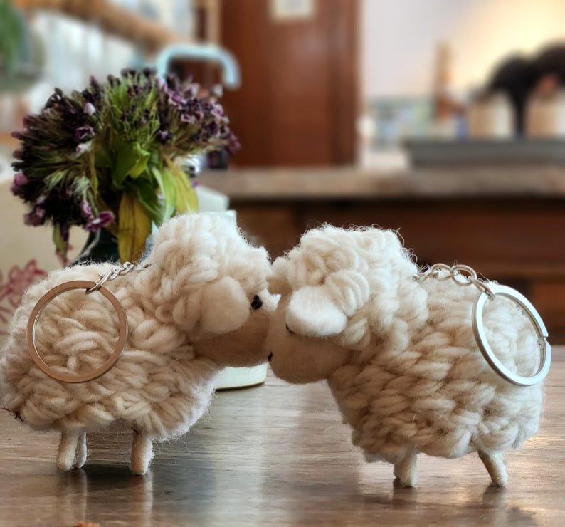 May include: Two off-white sheep keychains made of a textured, wool-like material. Each sheep has a silver-colored keyring attached. The sheep are positioned close together, suggesting a pair. The background is blurred, with hints of a floral arrangement.
