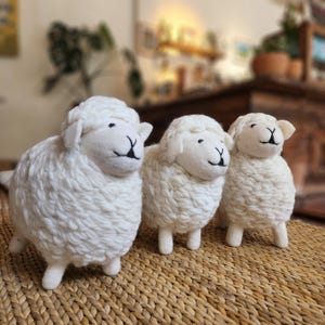May include: Three white stuffed sheep toys with smiling faces and black accents. The sheep have fluffy wool-like bodies and stand on four white legs. They are arranged on a woven, brown surface.