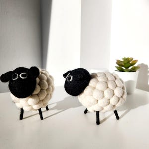 Puede incluir: Dos ovejas de fieltro blanco con caras y patas negras. Las ovejas están de pie sobre una superficie blanca.