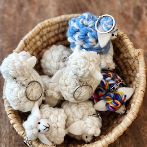 May include: A woven basket filled with handmade sheep keychains. The keychains are made of white and blue yarn, with silver key rings. Some sheep have colorful yarn accents. The basket sits on a wooden surface.