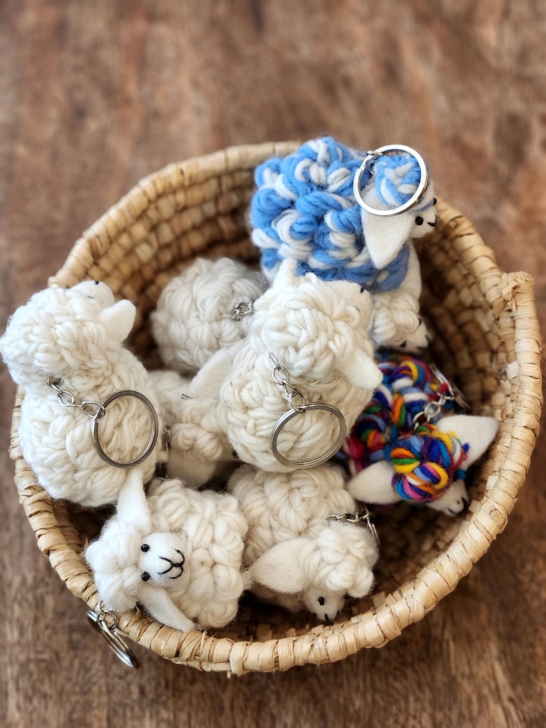 May include: A woven basket filled with handmade sheep keychains. The keychains are made of white and blue yarn, with silver key rings. Some sheep have colorful yarn accents. The basket sits on a wooden surface.