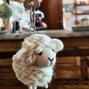 May include: A handmade, white sheep keychain with a silver key ring. The sheep is crafted from fluffy yarn, featuring black eyes and a black nose. The keychain is attached to a silver key with the word "Persian" on it.