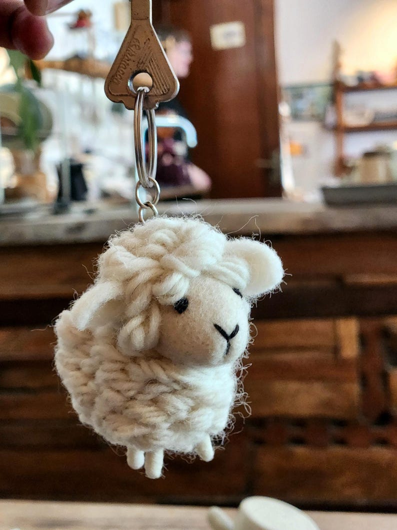May include: A handmade, white sheep keychain with a silver key ring. The sheep is crafted from fluffy yarn, featuring black eyes and a black nose. The keychain is attached to a silver key with the word "Persian" on it.