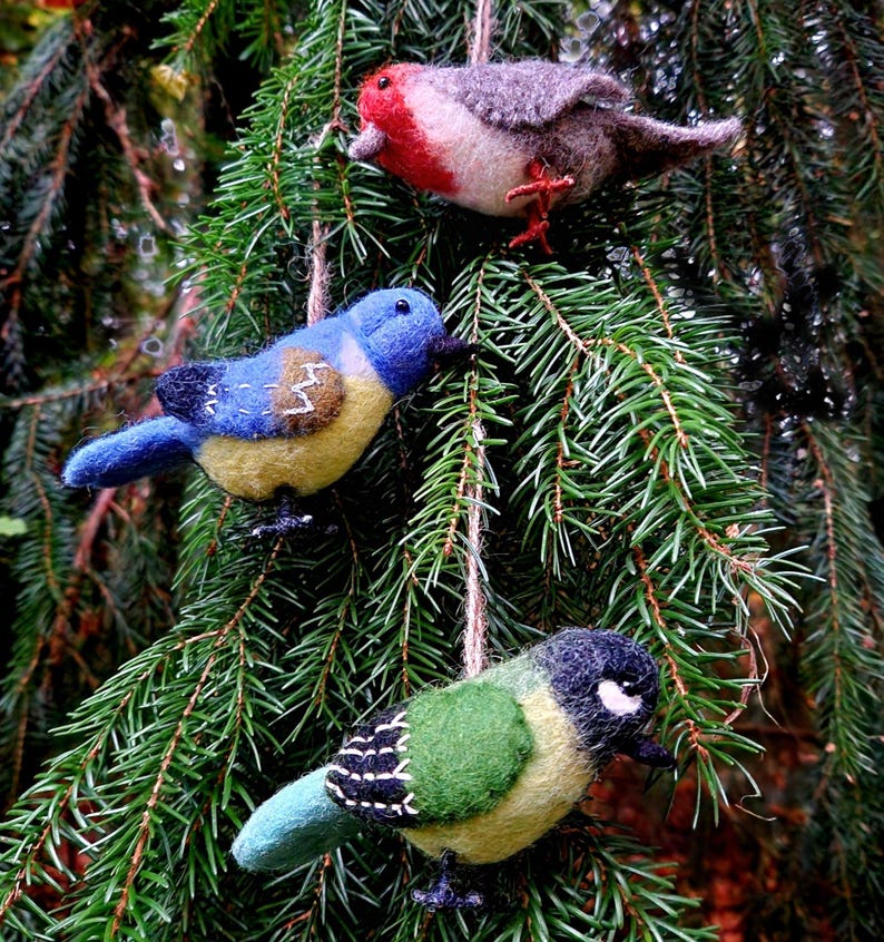 May include: Three felt bird ornaments hanging from a pine tree branch. The birds are blue, green, and red with yellow accents.