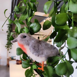 May include: A grey and white felt bird with a red breast perched on a green leafy plant. The bird has orange feet and a small beak.
