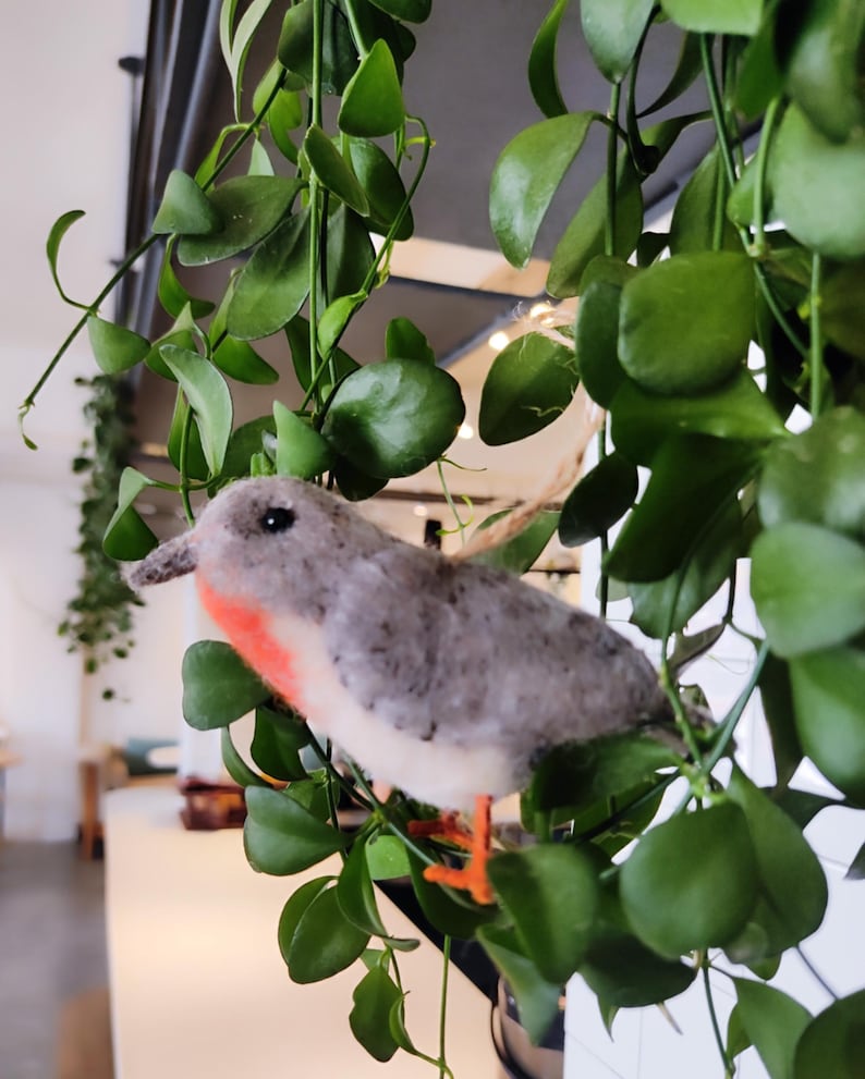 May include: A grey and white felt bird with a red breast perched on a green leafy plant. The bird has orange feet and a small beak.