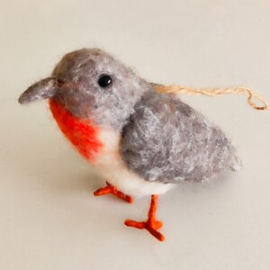 May include: A small, grey and white felt bird ornament with red accents and orange legs. The bird has a small, black eye and a string attached to the top of its head.