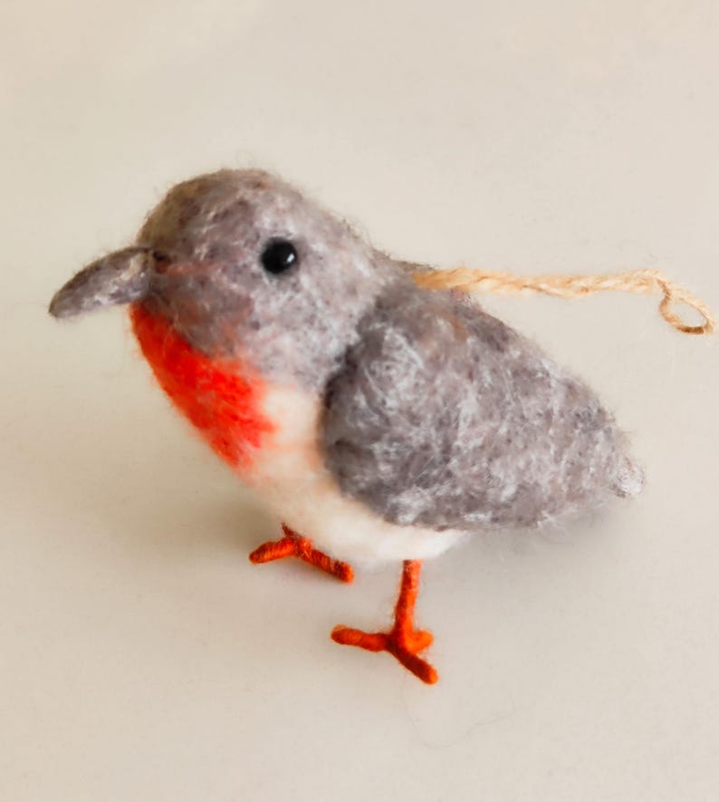 May include: A small, grey and white felt bird ornament with red accents and orange legs. The bird has a small, black eye and a string attached to the top of its head.