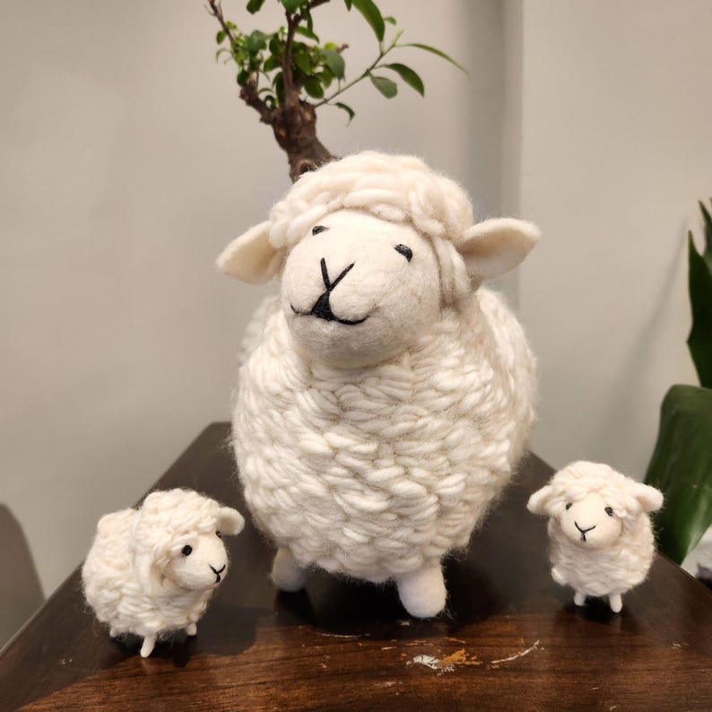 May include: Three white sheep figurines. The largest sheep is in the center, with two smaller sheep on either side. The sheep have fluffy wool-like textures, black eyes, and black facial details. The figurines are on a wooden surface.