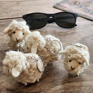 May include: Four handmade sheep keychains with silver keyrings. The sheep are made of white yarn and have black eyes and small legs. A pair of black sunglasses rests on a wooden surface.