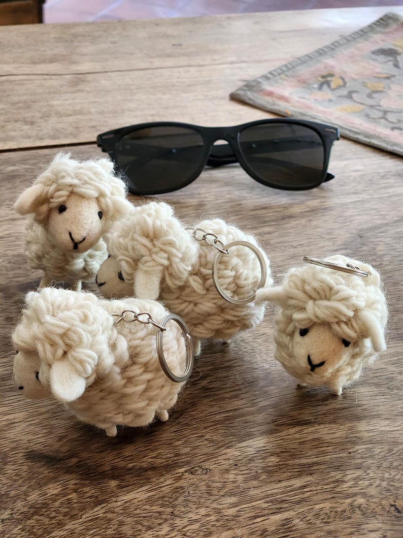 May include: Four handmade sheep keychains with silver keyrings. The sheep are made of white yarn and have black eyes and small legs. A pair of black sunglasses rests on a wooden surface.