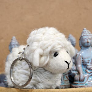 May include: A white, fluffy sheep keychain with a silver keyring. The sheep has black eyes and a black mouth. The background includes light blue Buddha statues and a tan surface.