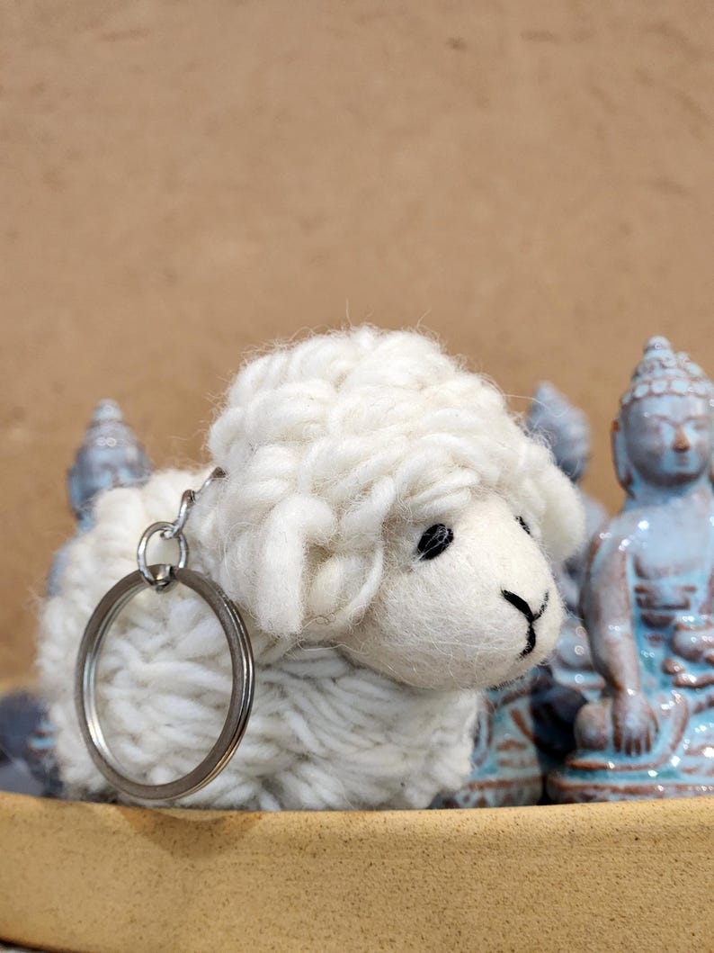 May include: A white, fluffy sheep keychain with a silver keyring. The sheep has black eyes and a black mouth. The background includes light blue Buddha statues and a tan surface.