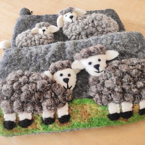 May include: A gray felted pouch featuring four three-dimensional sheep appliqu&eacute;s. Two sheep are positioned on a green grassy base at the bottom, while two smaller sheep are attached to the upper section. Each sheep has a white face and dark wool.