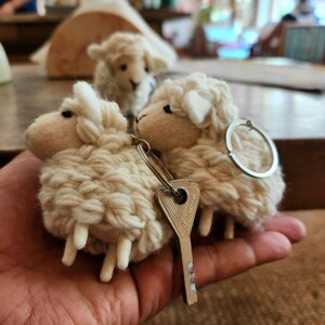 May include: Three woolly sheep keychains. The sheep are beige with white faces and legs. A silver key ring and a silver key are attached to two of the sheep. The keychains are held in a person's hand.
