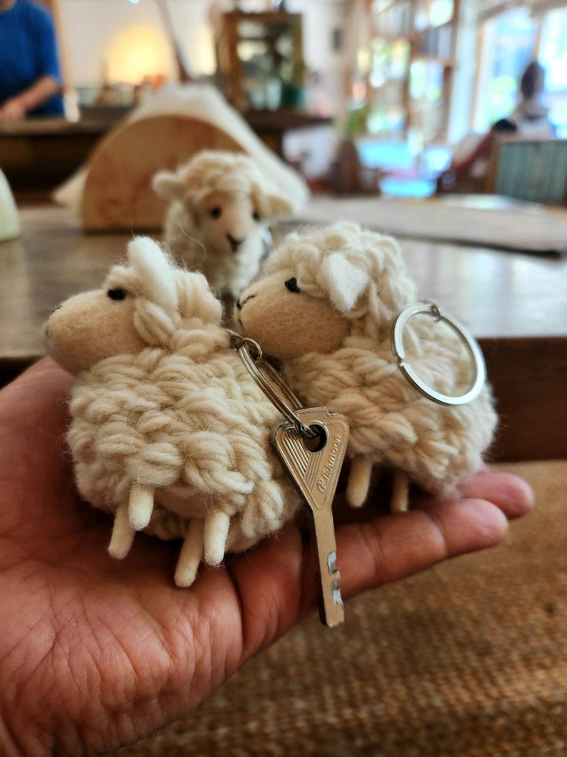 May include: Three woolly sheep keychains. The sheep are beige with white faces and legs. A silver key ring and a silver key are attached to two of the sheep. The keychains are held in a person's hand.