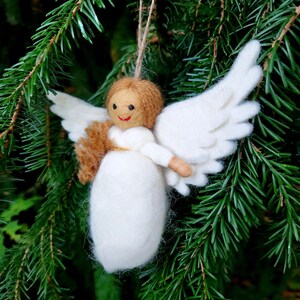 Puede incluir: Un adorno de &aacute;ngel de fieltro blanco con cabello casta&ntilde;o y un halo dorado. El &aacute;ngel sostiene un peque&ntilde;o objeto de fieltro marr&oacute;n en sus manos. El adorno est&aacute; colgado de una rama de un &aacute;rbol de hoja perenne verde.