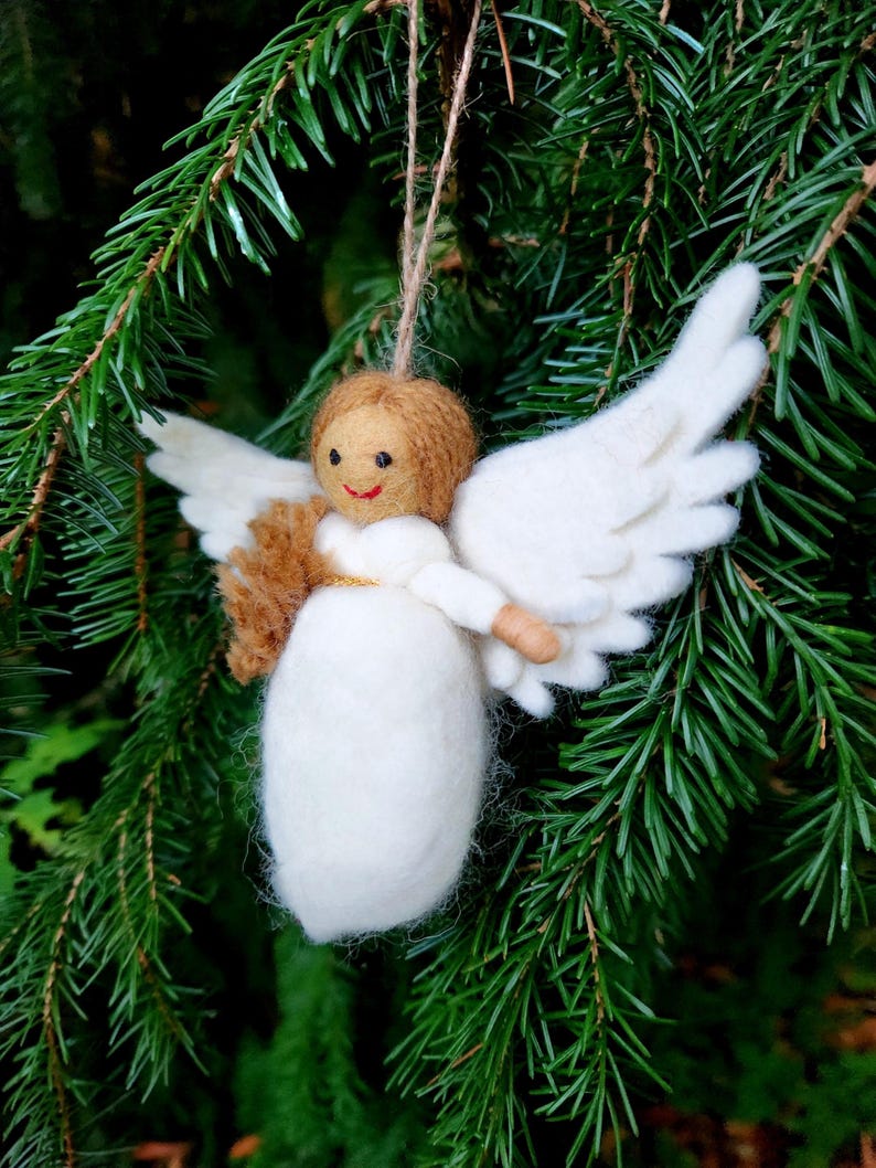 Puede incluir: Un adorno de &aacute;ngel de fieltro blanco con cabello casta&ntilde;o y un halo dorado. El &aacute;ngel sostiene un peque&ntilde;o objeto de fieltro marr&oacute;n en sus manos. El adorno est&aacute; colgado de una rama de un &aacute;rbol de hoja perenne verde.
