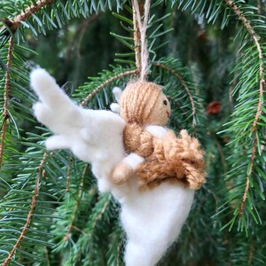 Puede incluir: Un adorno de &aacute;ngel de fieltro blanco con pelo y alas de lana marr&oacute;n. El &aacute;ngel est&aacute; colgado de una rama de un &aacute;rbol de hoja perenne verde.