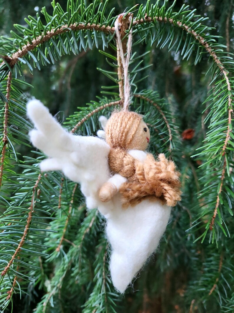 Puede incluir: Un adorno de &aacute;ngel de fieltro blanco con pelo y alas de lana marr&oacute;n. El &aacute;ngel est&aacute; colgado de una rama de un &aacute;rbol de hoja perenne verde.