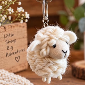 May include: A cream-coloured sheep keyring crafted from thick yarn, featuring a round body, small ears, and a simple face with black thread details for eyes and a nose. The sheep is attached to a silver-toned metal split ring for keys.
