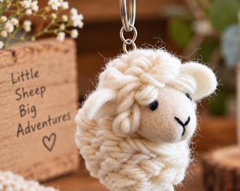 May include: A cream-colored sheep keychain crafted from thick yarn, featuring a round body, small ears, and a simple face with black thread details for eyes and a nose. The sheep is attached to a silver-toned metal split ring for keys.