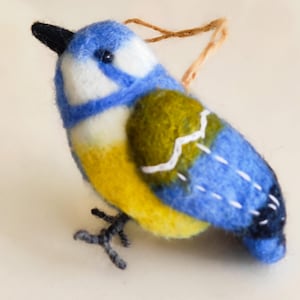 May include: A felt bluebird ornament with a yellow belly, green wings, and a black beak. The bird has a white stripe on its head and a white stripe on its wings. The ornament has a brown string for hanging.