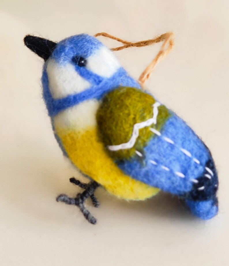 May include: A felt bluebird ornament with a yellow belly, green wings, and a black beak. The bird has a white stripe on its head and a white stripe on its wings. The ornament has a brown string for hanging.