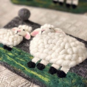 May include: A needle felted wall hanging featuring two white sheep with black hooves standing on a green textured field against a dark gray background. A small gray felt ball is attached to the top corner of the rectangular gray felt base.