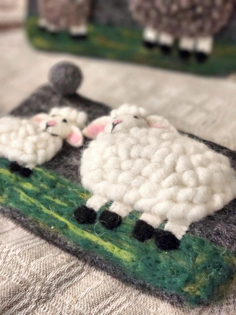 May include: A needle felted wall hanging featuring two white sheep with black hooves standing on a green textured field against a dark gray background. A small gray felt ball is attached to the top corner of the rectangular gray felt base.