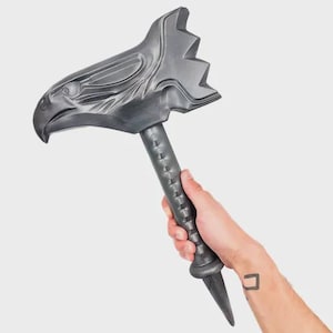 Pode incluir: Um martelo decorativo cinza escuro com um design de cabeça de águia. O martelo tem um cabo texturizado e uma extremidade pontiaguda. A cabeça da águia apresenta entalhes detalhados de penas e olhos. O martelo é segurado em uma mão contra um fundo branco.