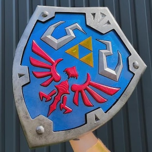 May include: A blue and silver shield with a red emblem and three yellow triangles. The shield has a silver border and is held up against a gray background. The shield is a prop from a video game.