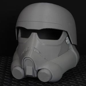 May include: A grey Stormtrooper helmet, a replica from a popular science fiction franchise. The helmet features a rounded top, a visor, and a detailed front with air vents and circular cheek pieces. The helmet is made of a smooth, matte material.