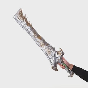 Op de afbeelding: Een zilveren en gouden fantasiezwaard met een gekartelde rand en decoratieve uitsparingen. De handgreep is zilver met groene accenten. Het zwaard wordt vastgehouden door een persoon met een zwarte mouw.